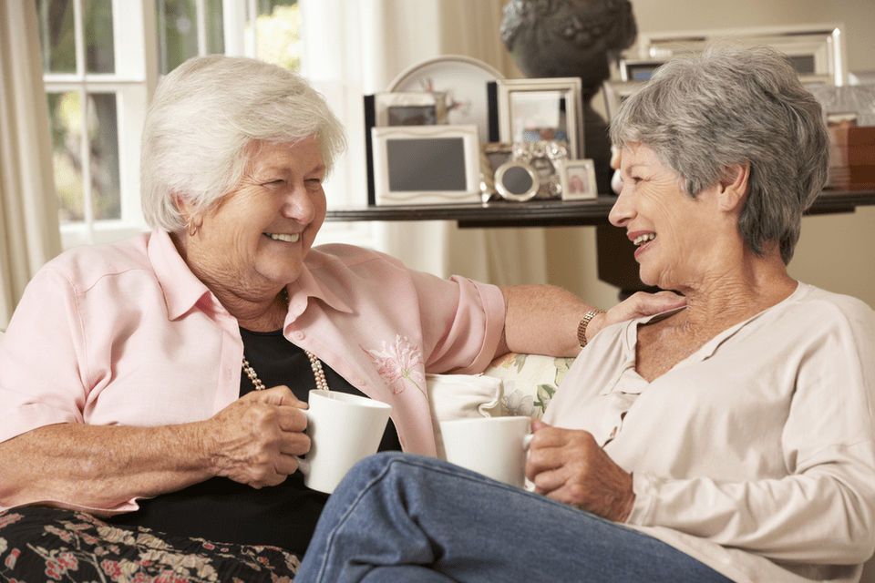 Two senior women sip coffee while sitting on a couch.
