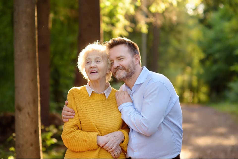 A senior woman and middle aged man hug while walking in a park.