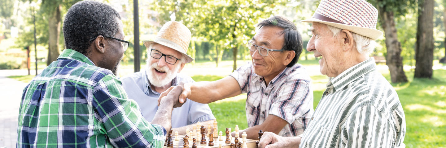 A group of four senior men smiling and laughing while playing chess in a park
