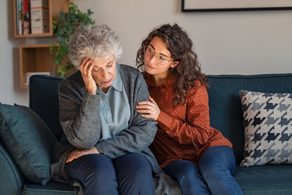 A woman comforts a senior woman while sitting on a couch.