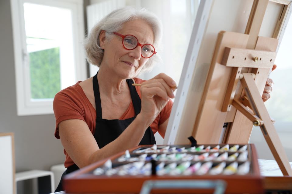 A senior woman in glasses paints at an easel.