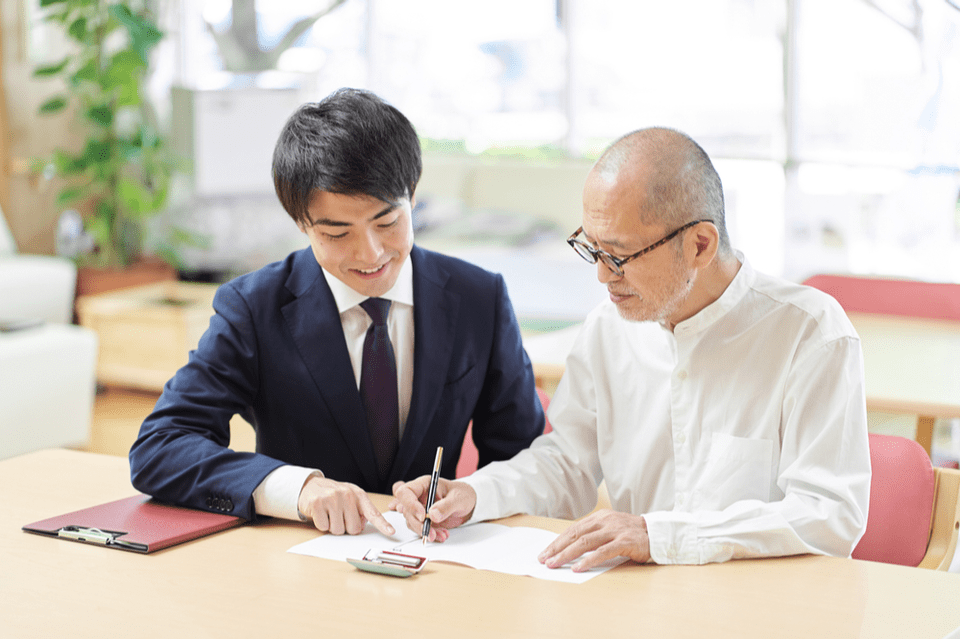 A senior man sits with a young man in a suit while signing a piece of paper.