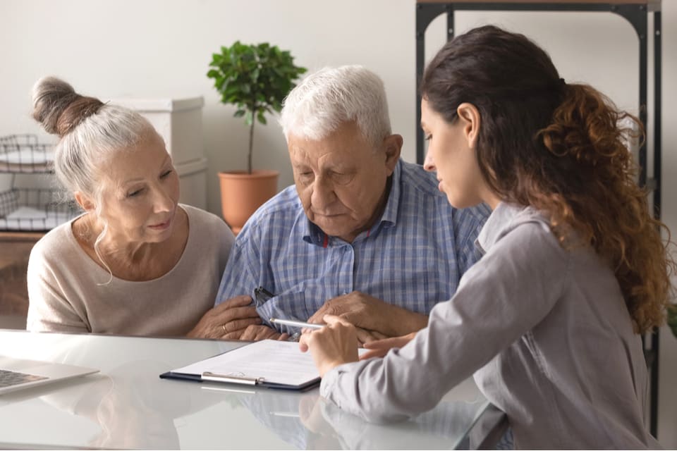 A senior man and woman sit at a table with a woman while looking at a contract.