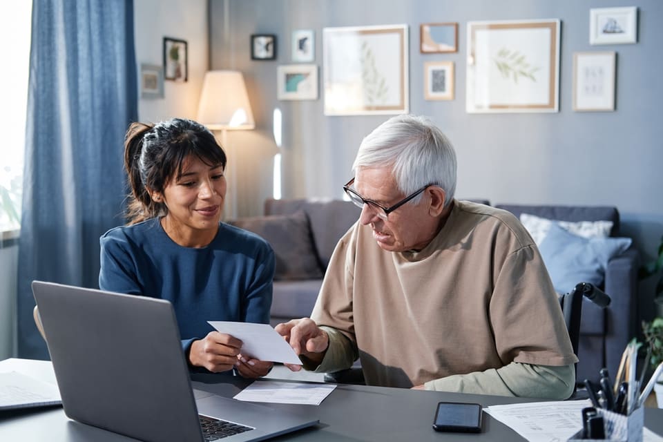 A senior man sits with a woman while looking at papers.