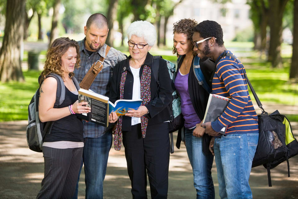 Four college students standing in a group showing page of a text book to a white haired senior woman.