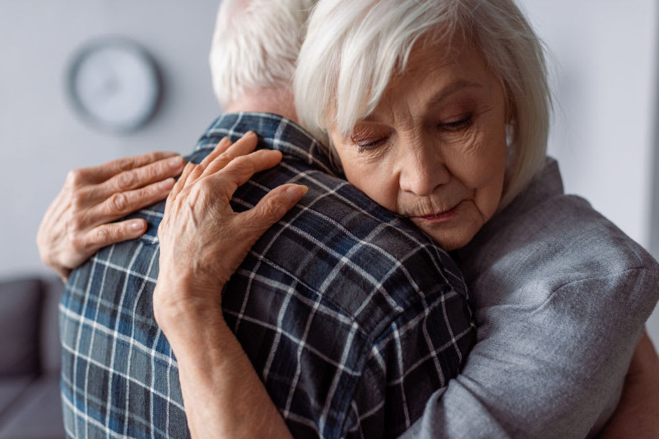 A senior woman hugs her spouse as they care for a parent with dementia and anger.