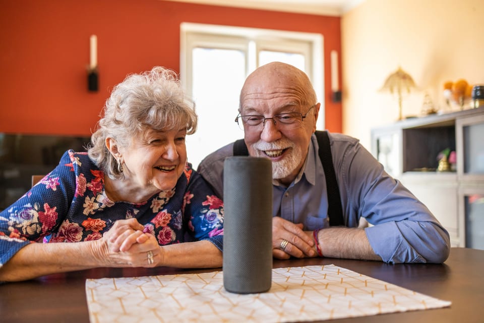 Two seniors sit at a table and talk to their Amazon device.
