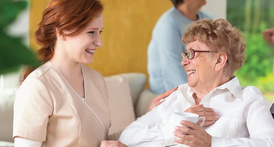 Older woman with glasses is seated, smiling at a younger woman with red hair. The younger woman is also smiling at the older woman, dressed in scrubs.