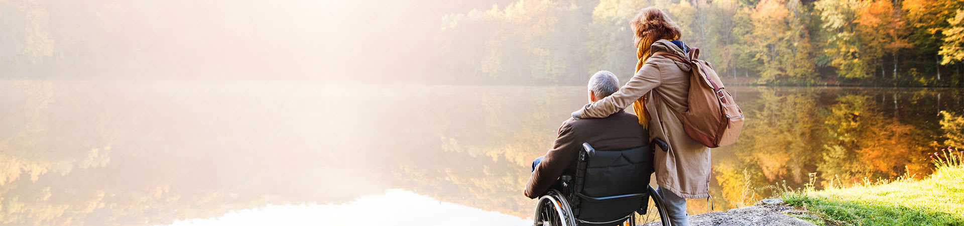 A view from behind of a woman with her arm around a senior man in a wheelchair while looking out at a serene pond