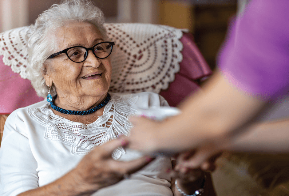 Senior woman sits in chair while being helped by caregiver.
