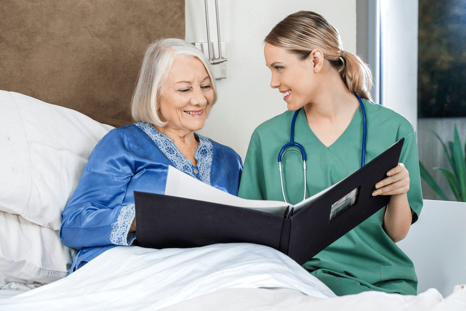 Young woman caregiver wearing stethescope sits on the site of a bed looking at an album with a smiling elderly woman.