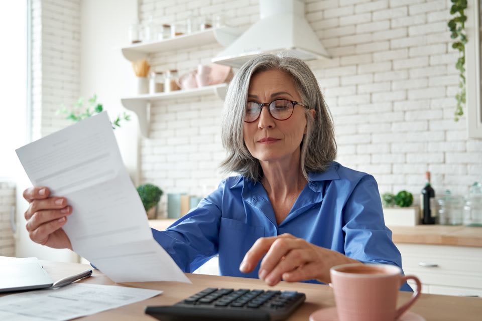 A senior woman sits at a table while typing on a calculator.