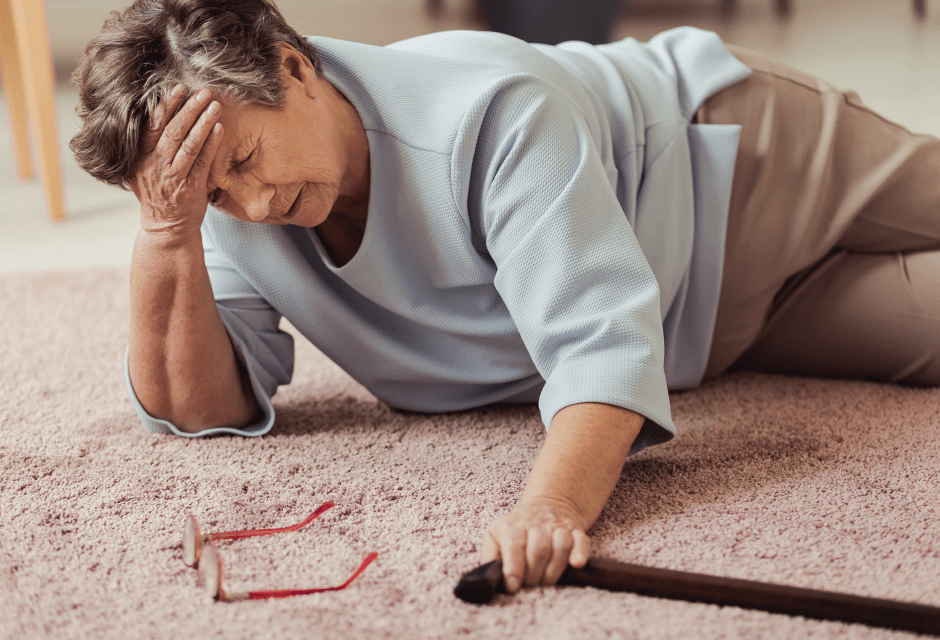 An elderly woman lies on a carpet, appearing distressed and holding her head with one hand, while her glasses and a cane are nearby.