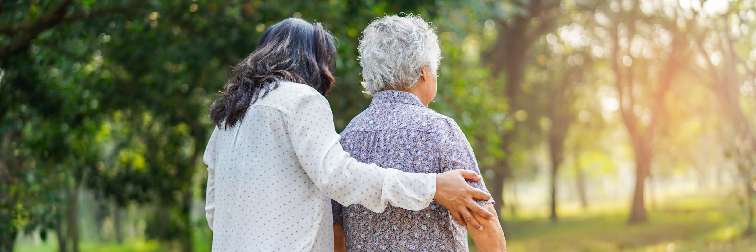 A view from behind of a home health care aide walking outdoors with a senior