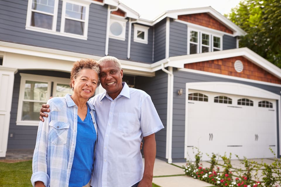 A man and woman smile while standing outside of a blue house.