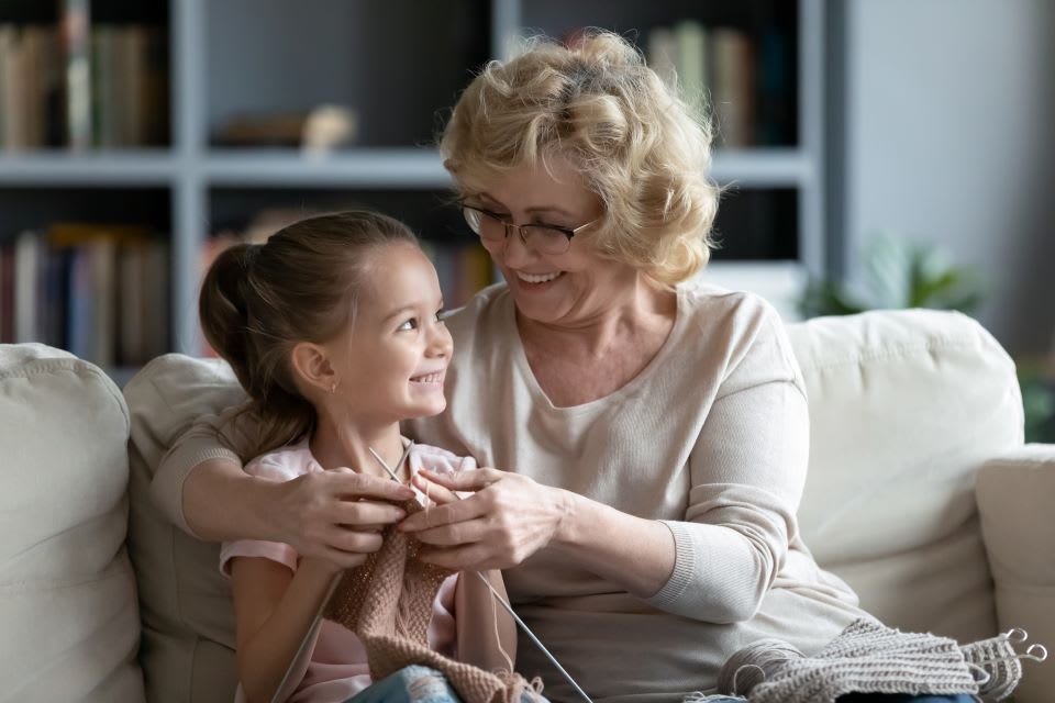 A senior woman and little girl sit side by side on a couch while smiling.