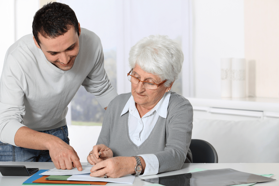 Senior woman looking at paperwork on desk while young man leans over her shoulder.