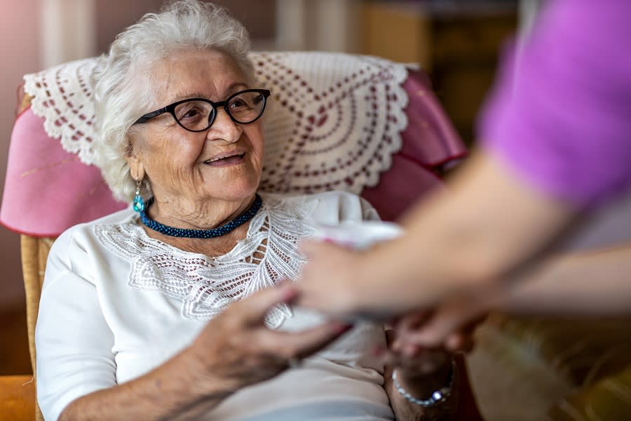A senior woman sits in a chair while holding hands with someone.