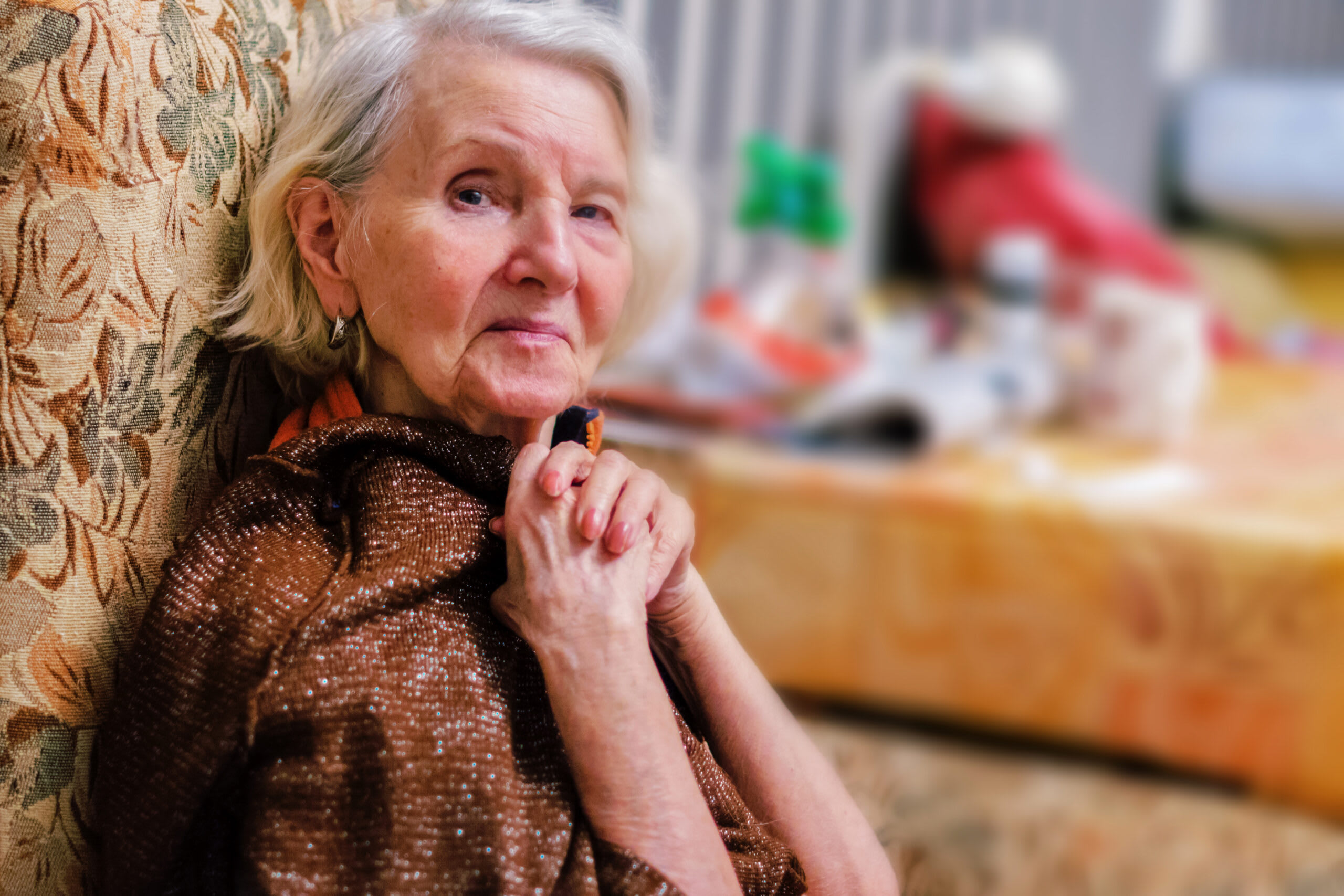 A senior woman sits in a chair with her hands clasped.