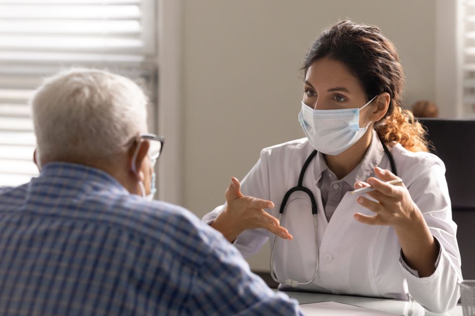 A senior man sits at a doctor's desk as they talk.