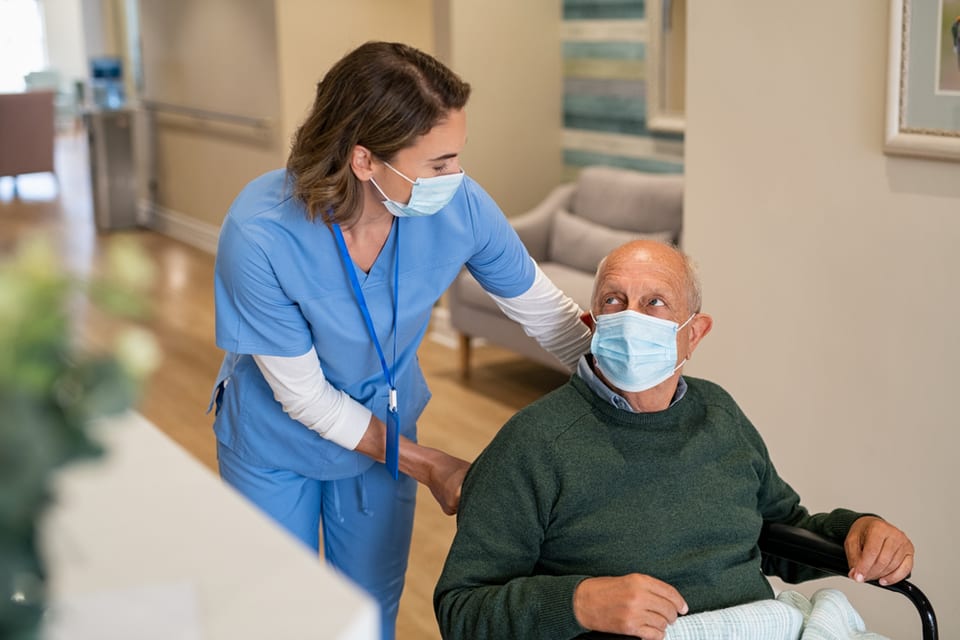 Masked caregiver pushes mask wearing older man in wheelchair.