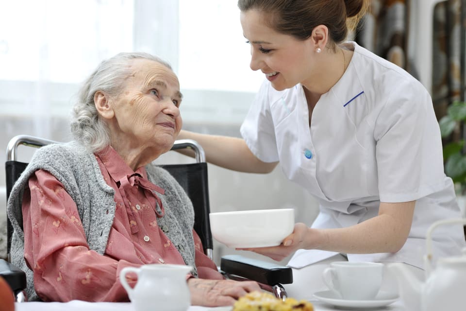 A senior woman sits at a table with food while a nurse assists her.