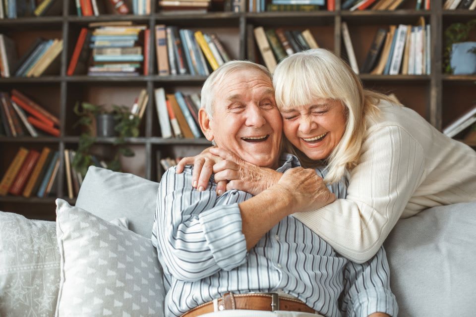 A senior man sits on the couch while hugging and laughing a woman.