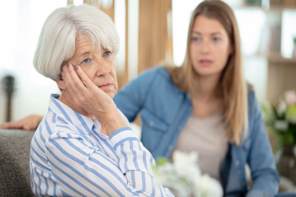 A senior woman expresses agitation while her caregiver looks on with concern.