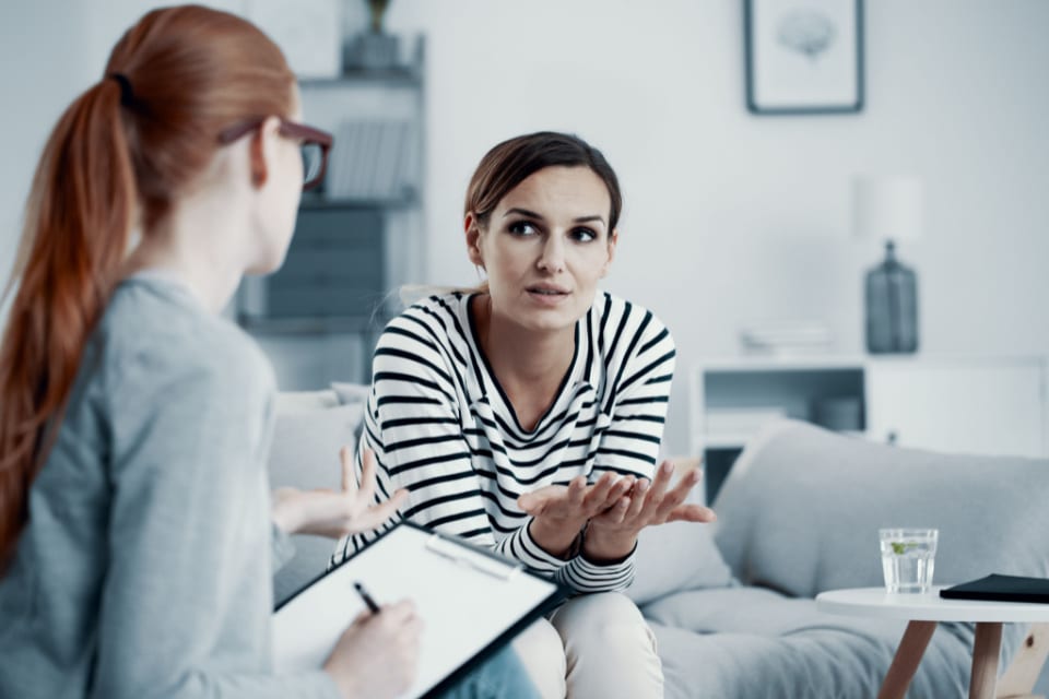 A woman sits on a couch in conversation with another woman who is taking notes