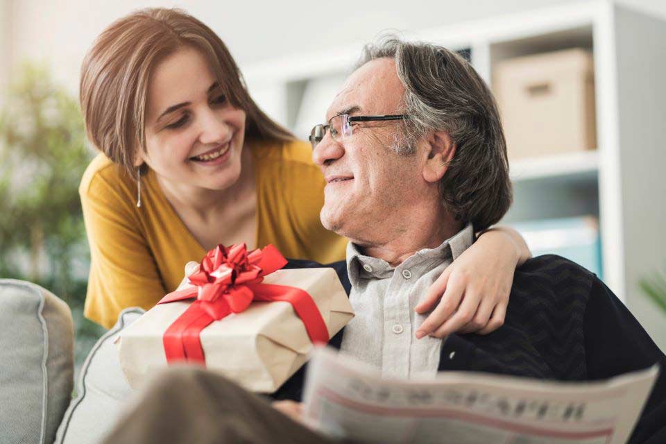 Young woman surprising her grandfather with a gift while he reads the newspaper.