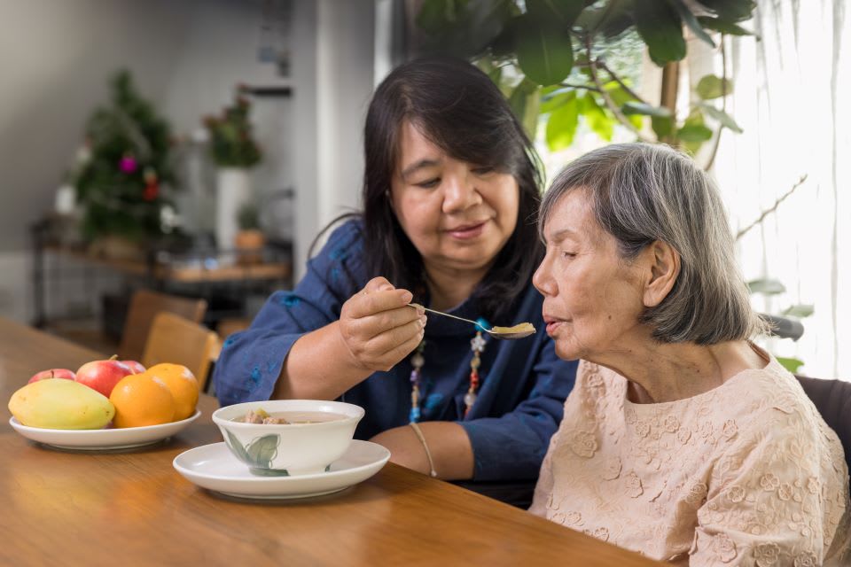 A home caregiver helps a senior woman to eat soup