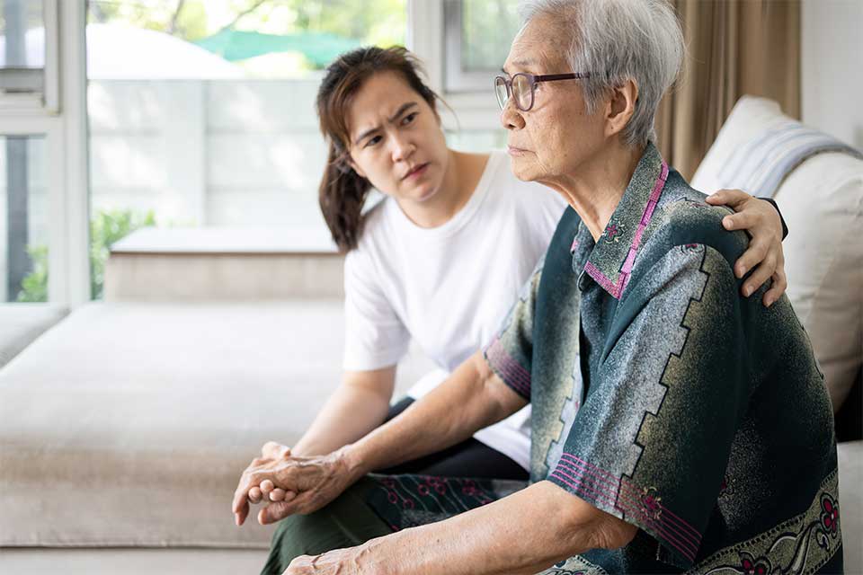 Elderly woman and her caregiver sitting on a couch trying to offer emotional support.