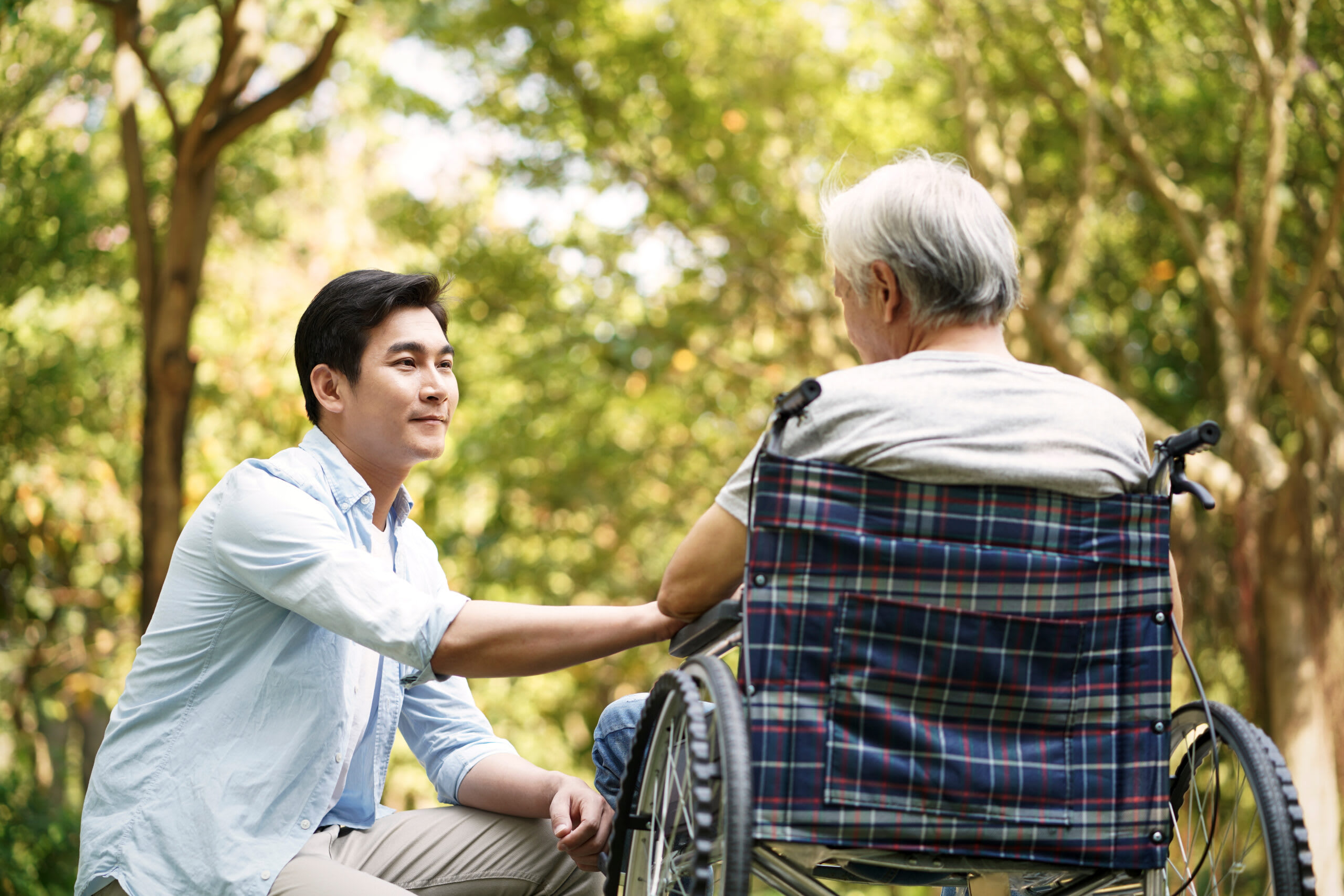 A man kneels and holds the hand of a senior man.