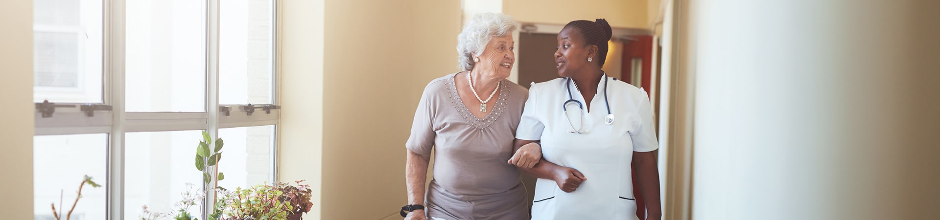 A nursing home caregiver helping a senior woman with a cane to walk down a hallway