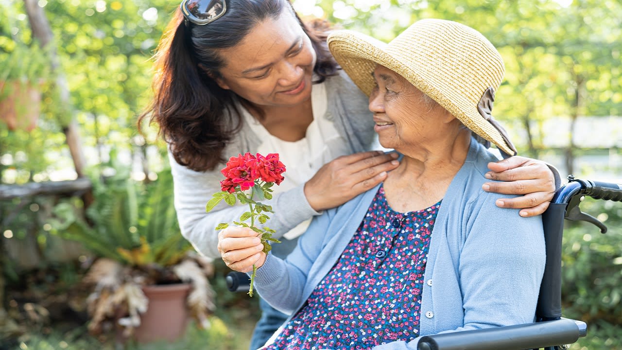 A senior woman and her caregiver smile while sitting in a garden.