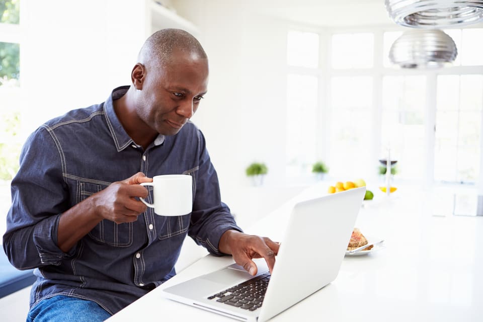 Older black man drinking coffee while researching grants for dementia patients on a laptop.