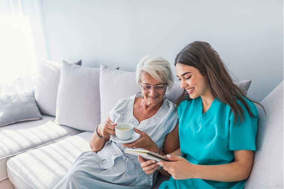 Elderly woman being read to by her private caregiver while sitting on a couch.