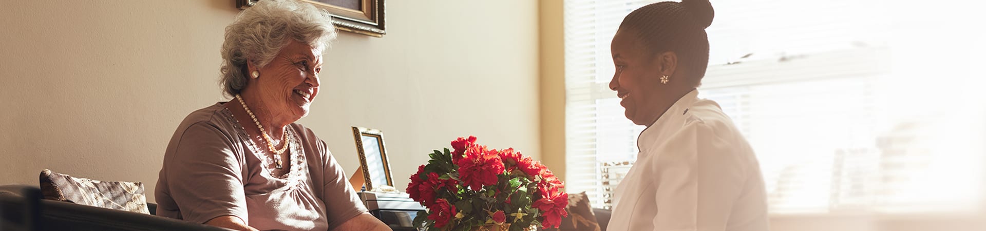 A senior and a caregiver smile at eachother as they sit in front of a bushel of red roses