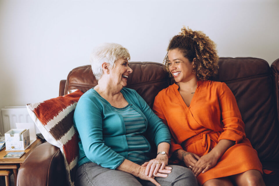 A senior woman and a younger woman sitting on a couch while smiling in conversation