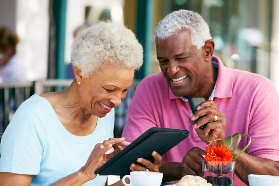 A senior man and woman laugh while using a tablet at a table.