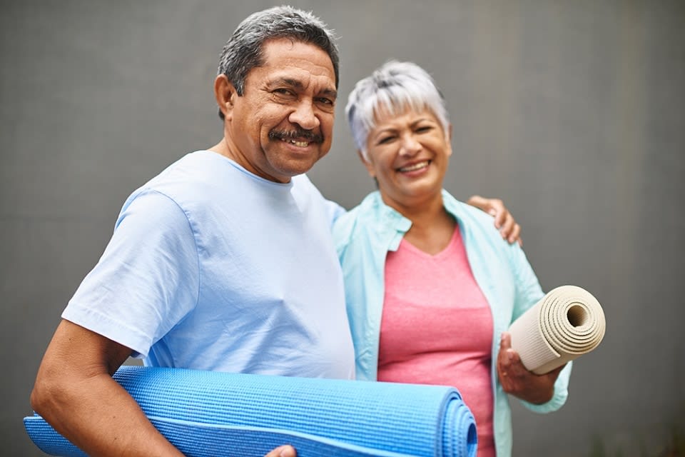Older man with his arm around older woman's shoulder, both are carrying yoga mats.
