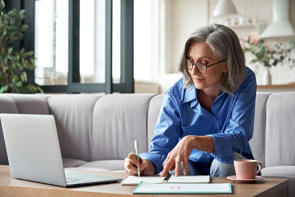 Grey haired middle age woman writing on tablet while looking at laptop screen.