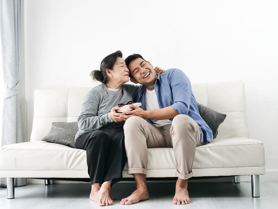 Mother kissing son on cheek while seated on couch with both of them holding a wrapped gift.