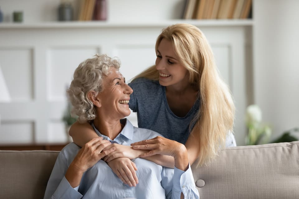 A senior woman and younger woman hug while sitting on the couch.