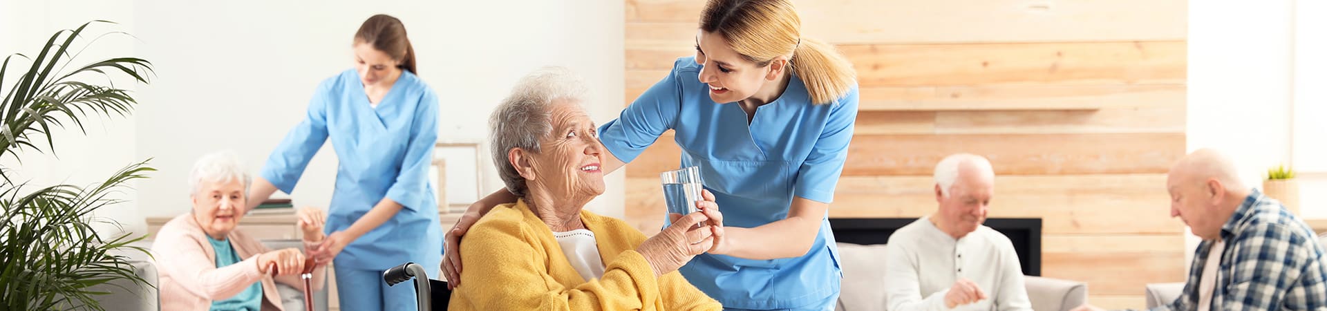 A group of seniors sitting in a common area while two nurses assist two senior women