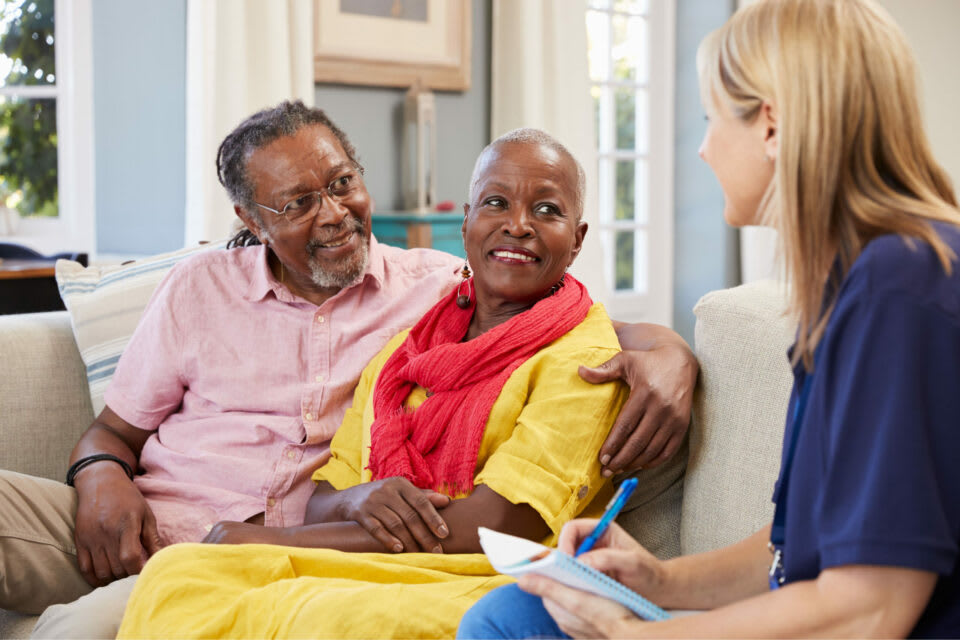 A caregiver sits on a couch in conversation with a senior couple while taking notes