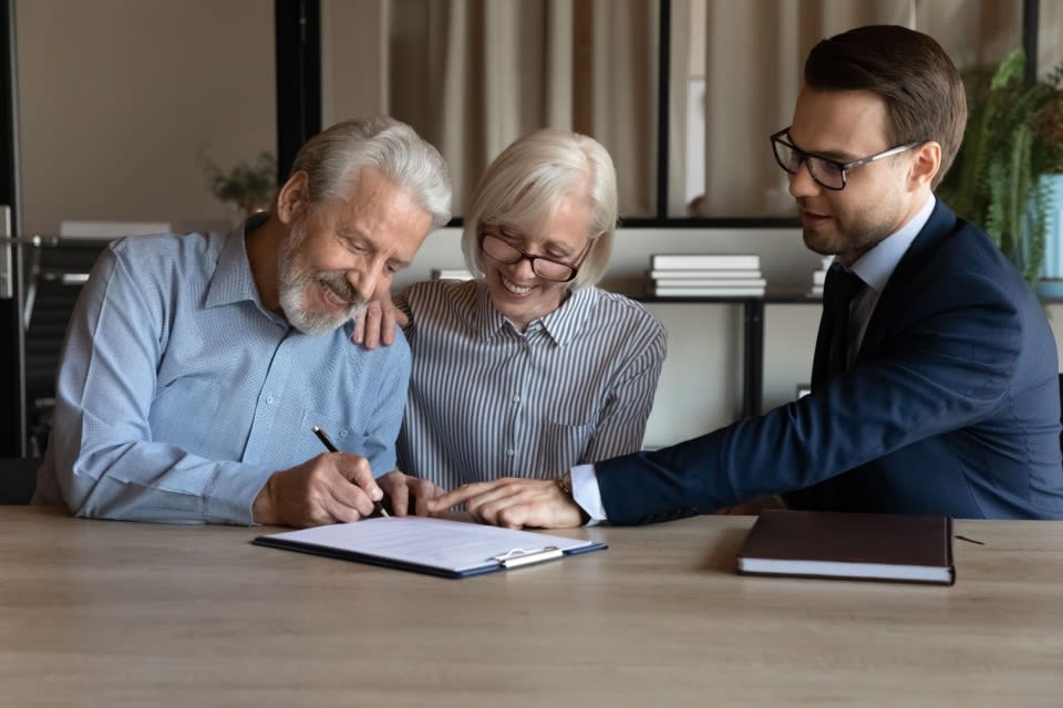 Smiling older couple sign legal documents as their attorney points to the signature lines