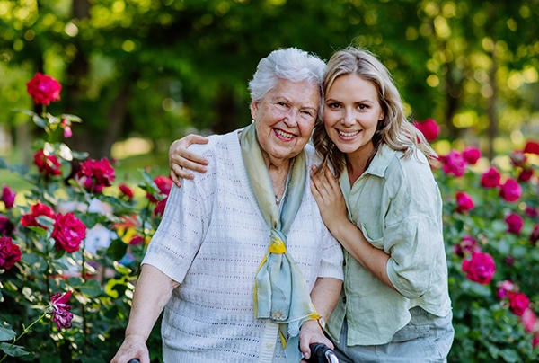 A middle-aged woman embracing her senior mother while they smile in front of rose bushes