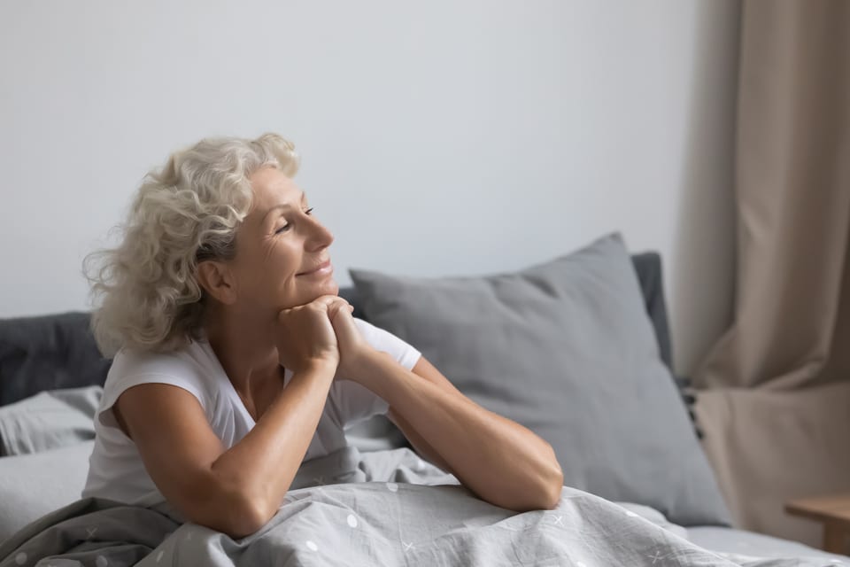 A senior woman sits in bed while smiling and looking out a window.