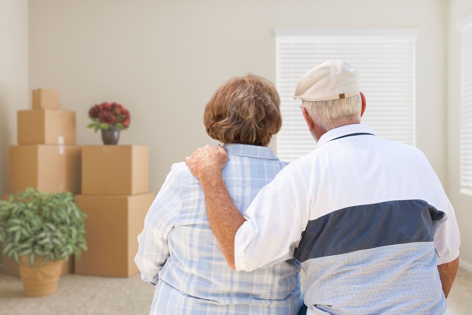 An elderly couple stands in a house filed with packed boxes.